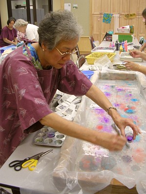 Gray-haired woman in purple brushes purple paint onto surface of the fluid in a marbling tray