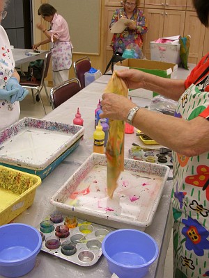 Hands raise dripping yellow cloth from marbling tray.