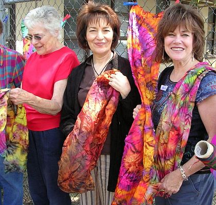 Three women hold up shibori fabric by a chainlink fence